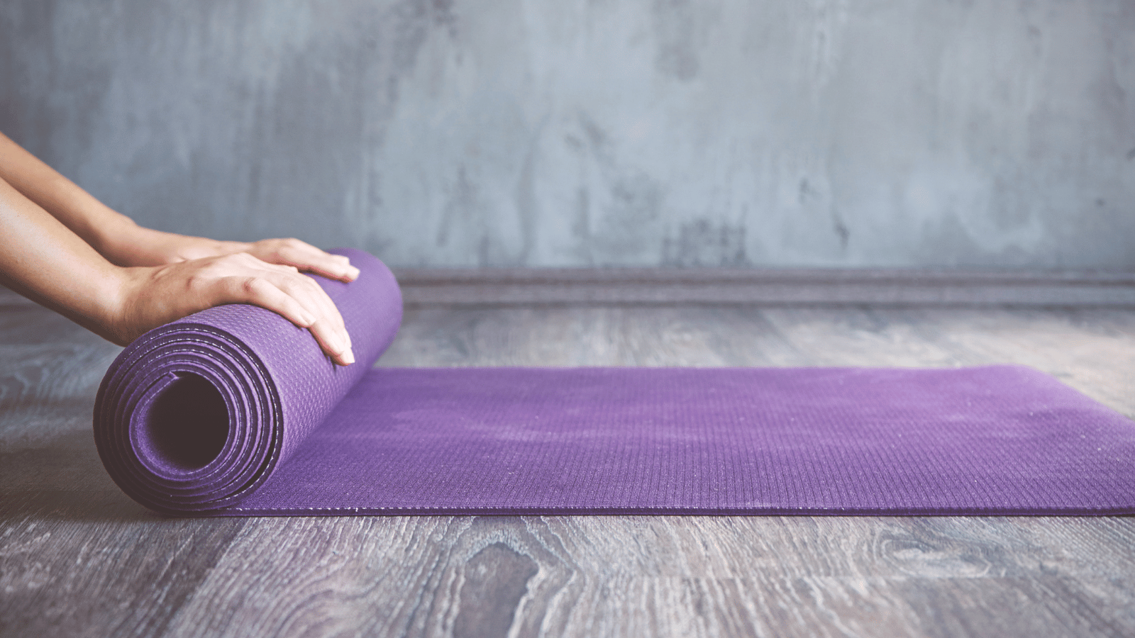 Woman's hands rolling out a purple yoga mat on a wooden floor against a gray concrete wall.